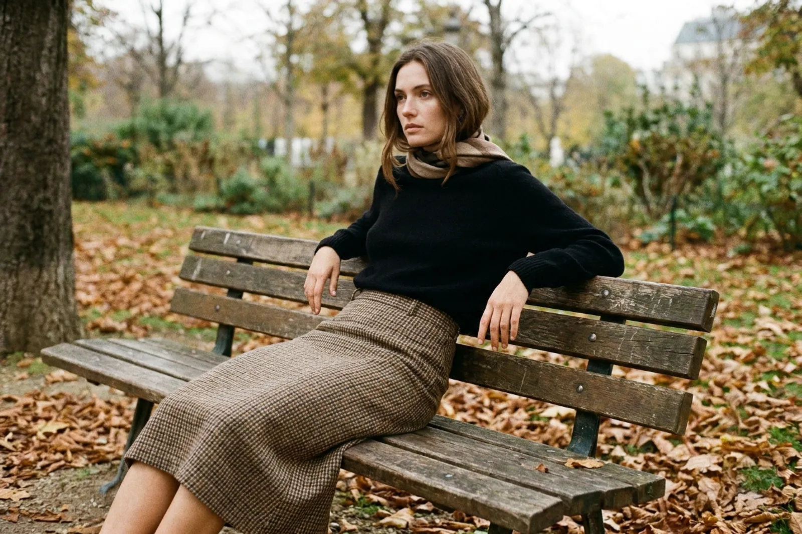 Une femme en jupe à carreaux et maille noire unie assise sur un banc, feuilles d'automne