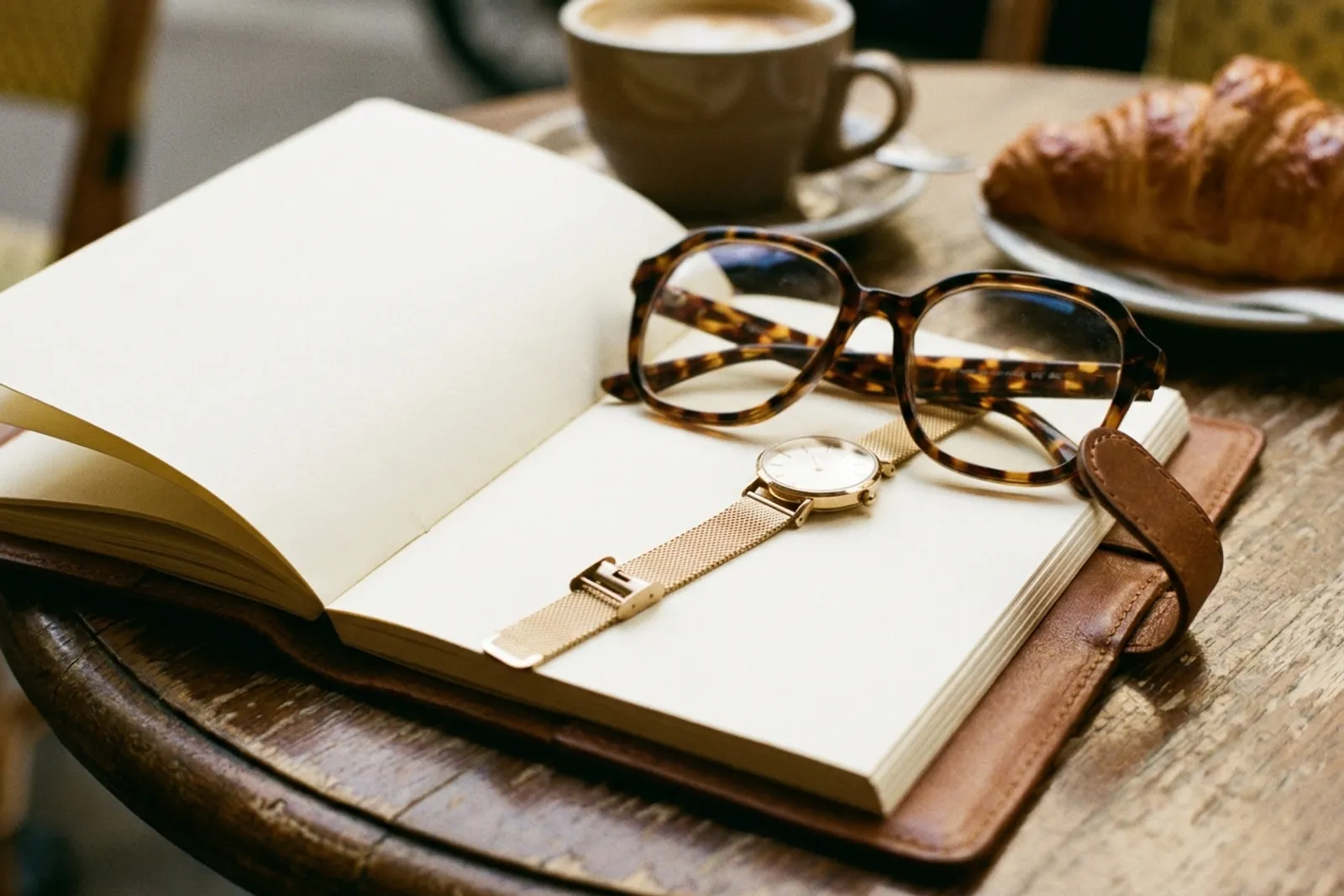 Nature morte de lunettes en écaille et d'une montre dorée fine posées sur un carnet ouvert, sur une table de café en lumière chaude